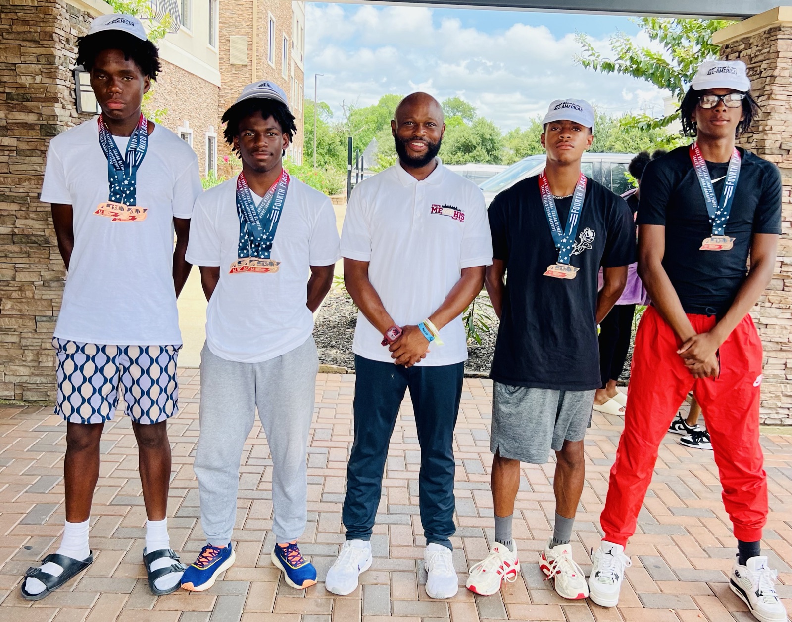 Coach Williams, in a white Youth Memphis Track & Field polo, stands at a hotel entrance between four teenage athletes, each wearing medals on star-patterned lanyards.