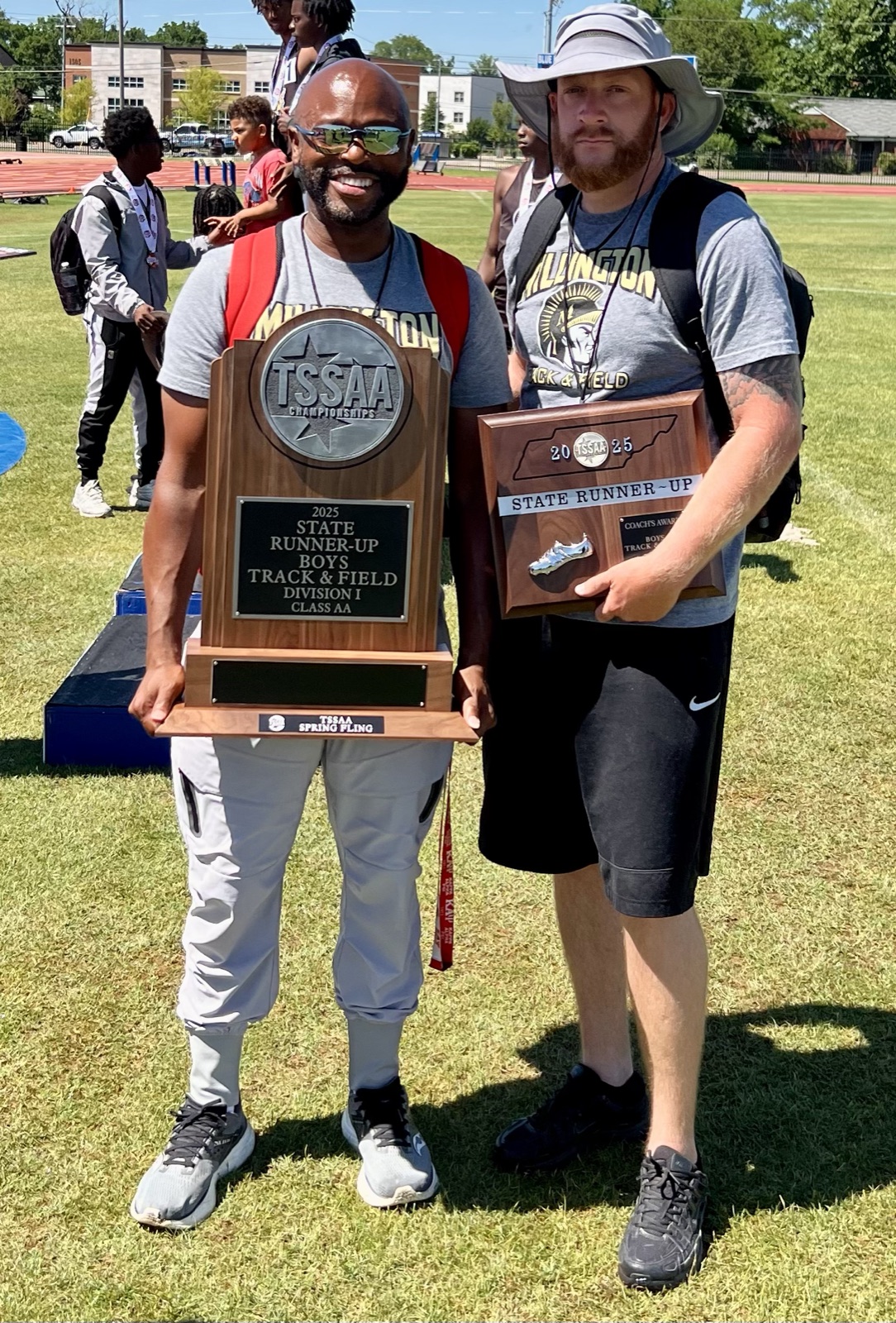 Coach Williams, in sunglasses and a Millington shirt, holds the wooden TSSAA state runner-up trophy on a track infield, beside a second coach holding a state runner-up plaque.
