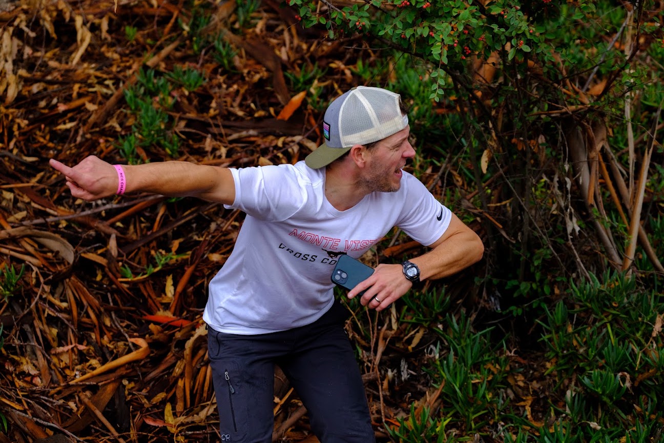 Coach Wales-Dinan in a cap, pointing the way on a cross country course.