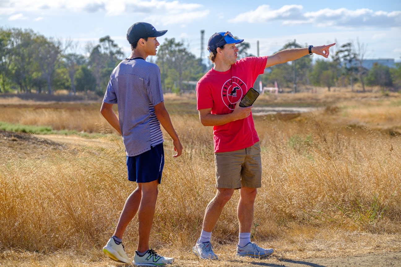 Coach Wales-Dinan, in a red Monte Vista shirt with his phone in one hand, points across a field of dry grass while a runner in a striped shirt looks the same way.